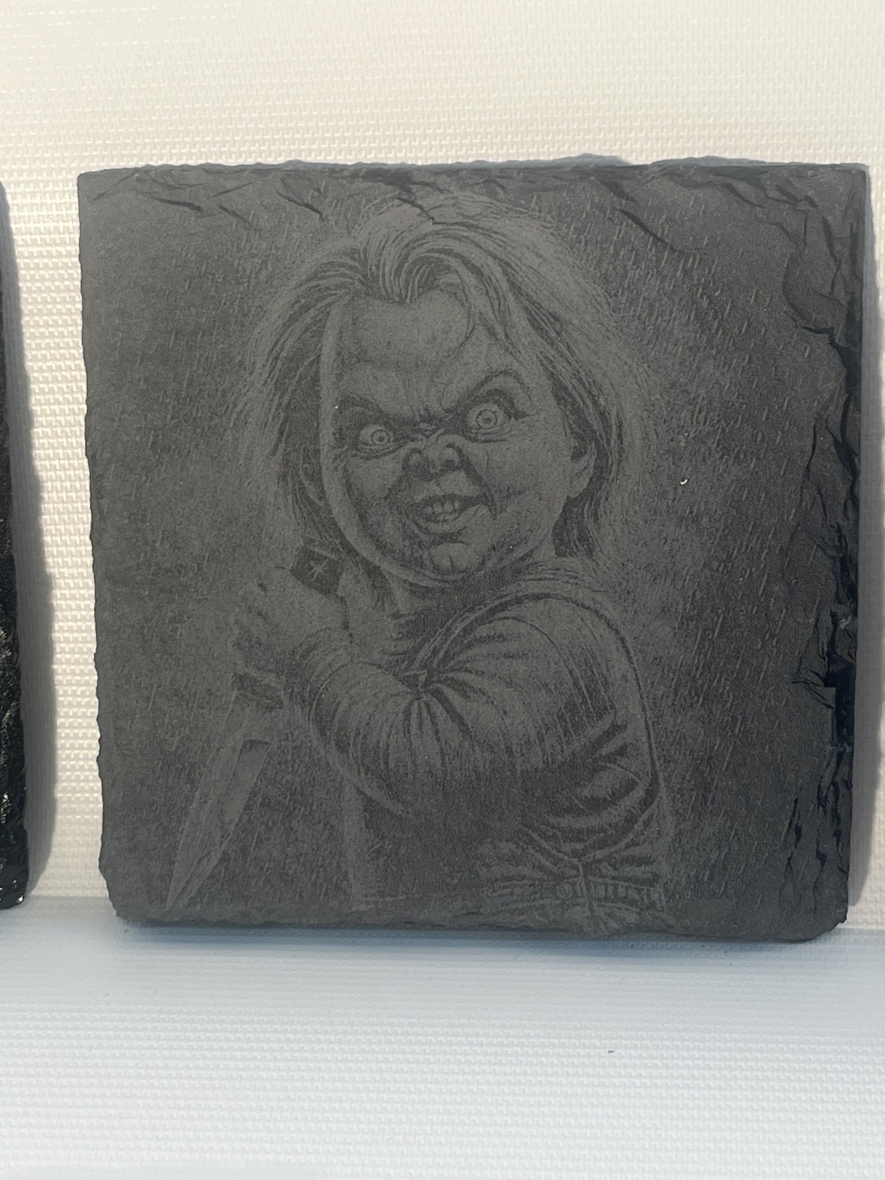 A square dark gray slate coaster featuring an embossed portrait of Chucky.