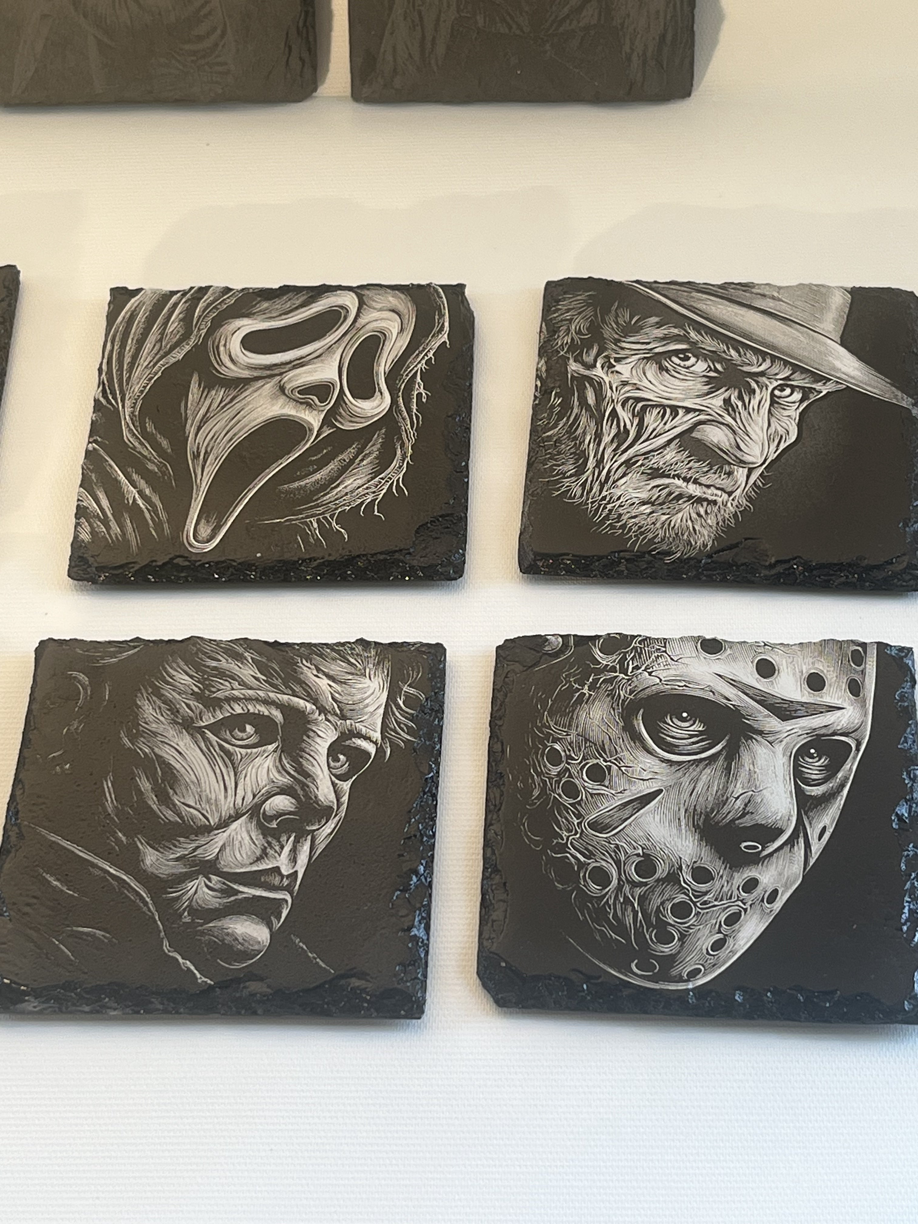 Four square dark slate coasters featuring detailed black and white carvings of horror movie faces, including Ghostface, Freddy Krueger, Michael Myers, and Jason Voorhees.