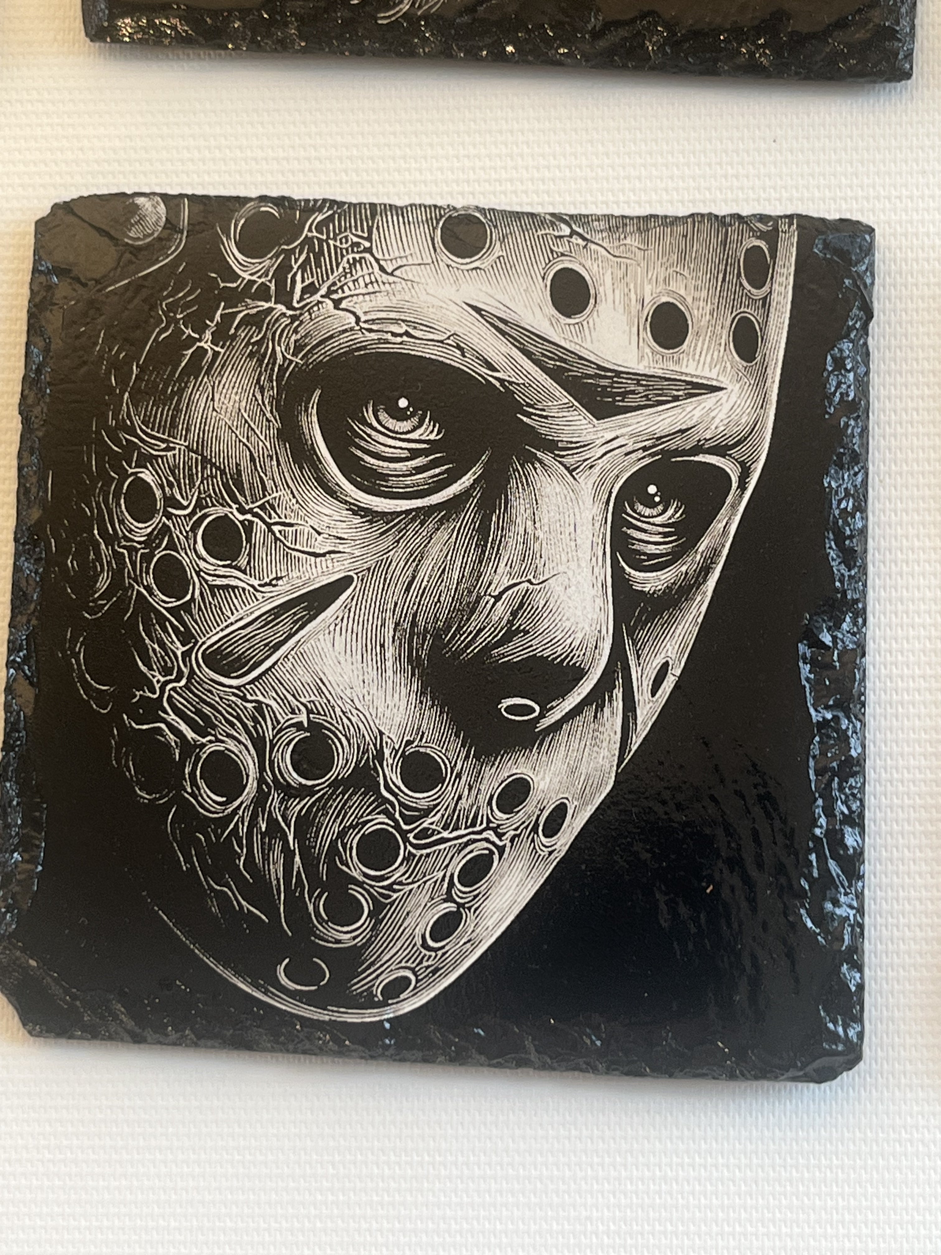 A square black slate coaster featuring a highly detailed black and white graphic illustration of a hockey mask.