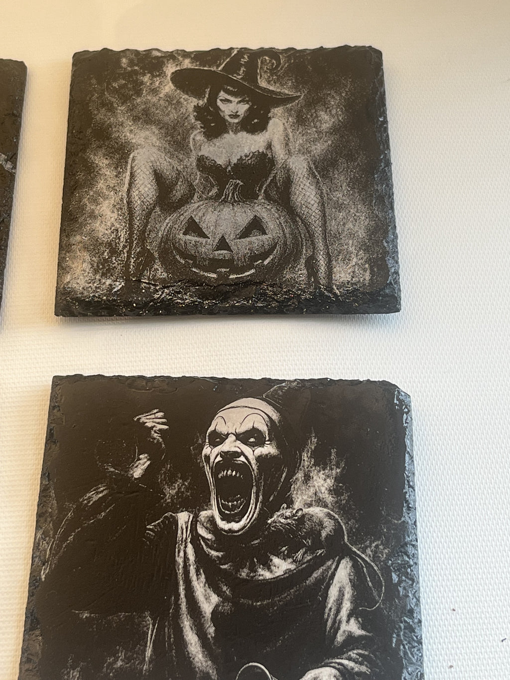 Two square black slate coasters featuring Halloween-themed designs, displayed on a white textured surface. The top coaster shows a witch sitting behind a carved jack-o'-lantern pumpkin.