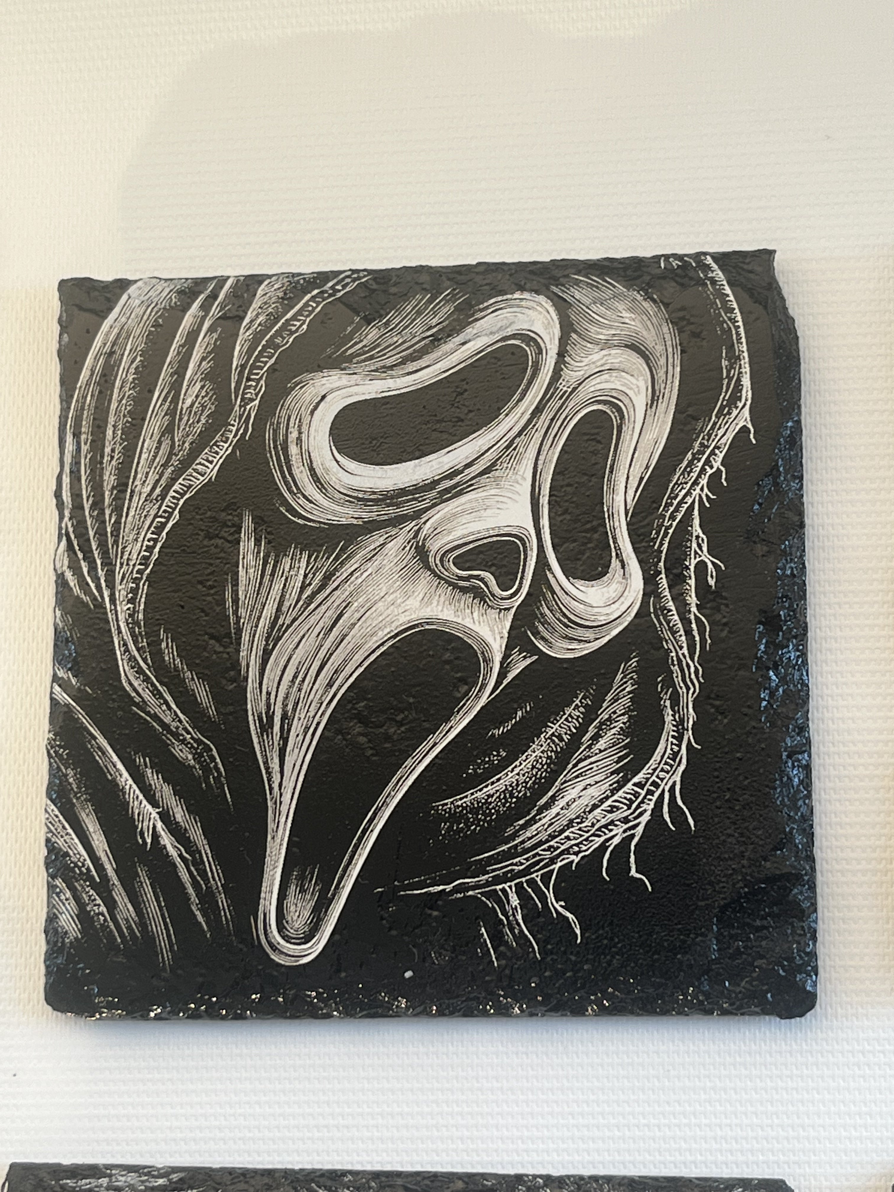 A square black slate coaster featuring a white and gray relief carving of the iconic Ghostface mask from the Scream franchise.