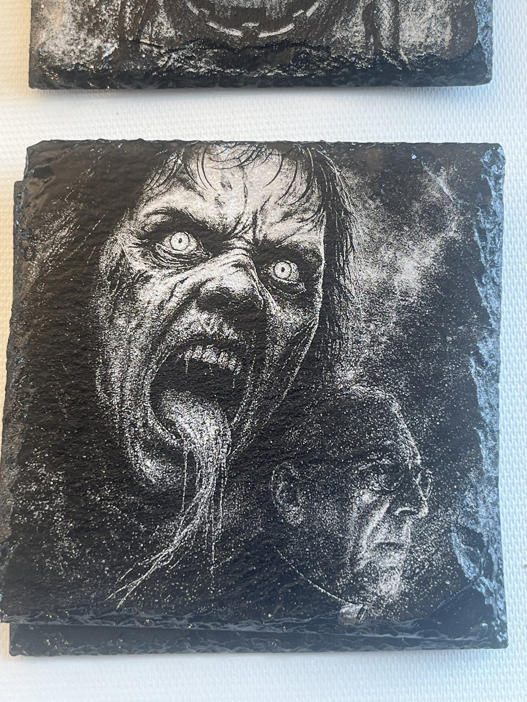 A square dark slate coaster featuring a highly detailed, high-contrast black and white relief carving of a horror character's screaming face, with a smaller profile view in the lower right.